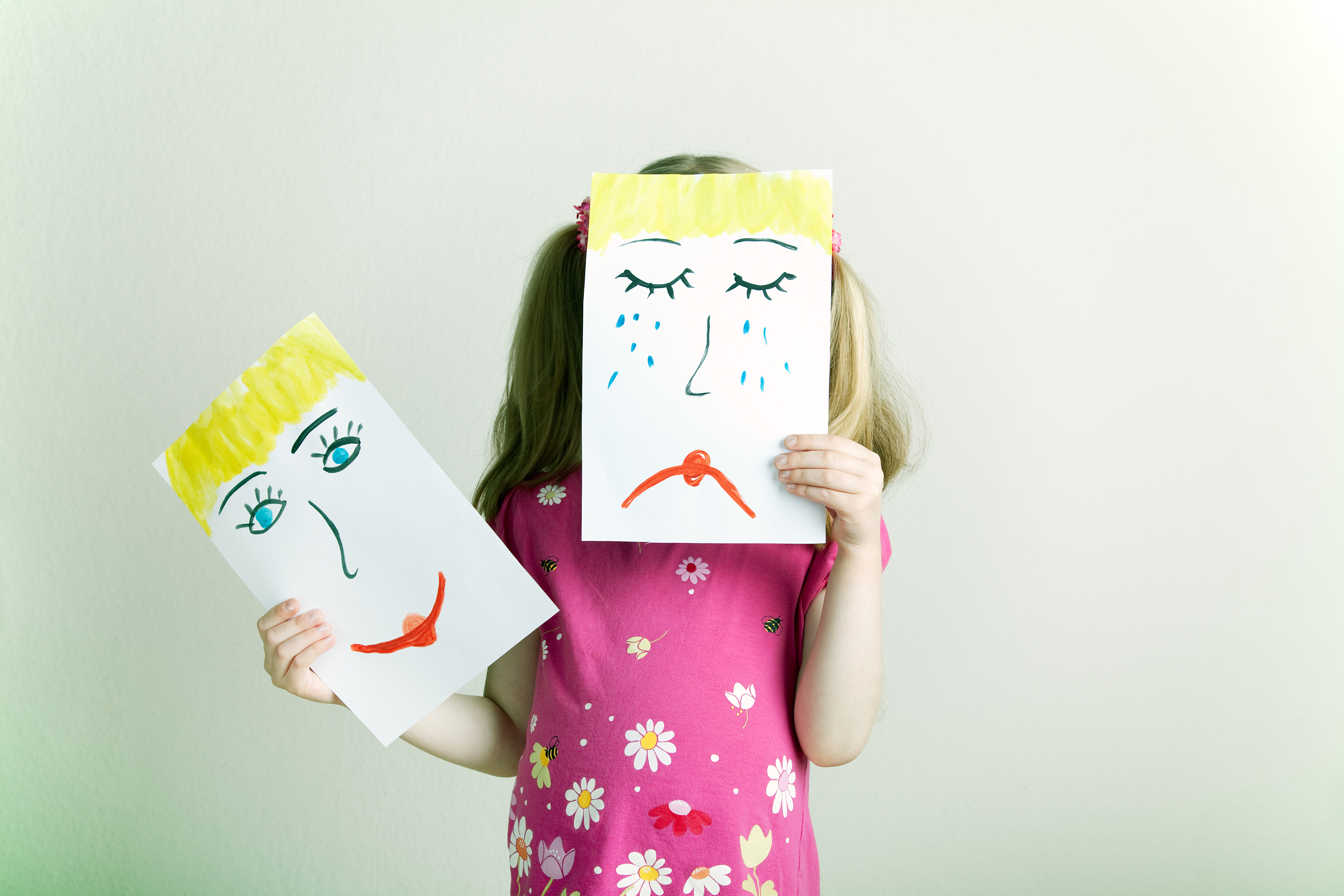 Child in pink floral shirt holding a drawing of a crying face in front of their own face and a drawing of a happy face in their free hand.