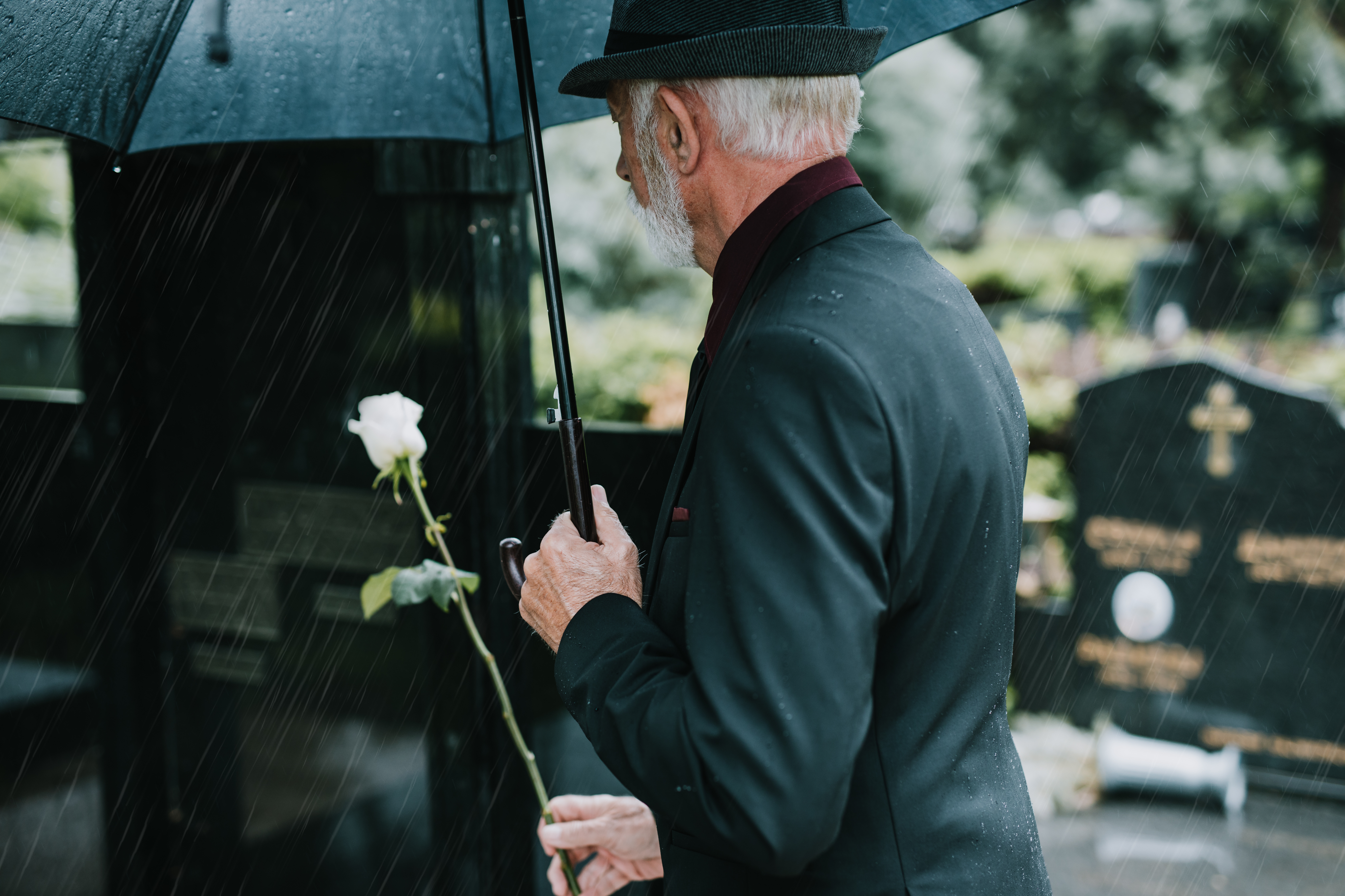 Man with white hair and beard, wearing black suit and hat, holding black umbrella and white flower, standing near gravestones.