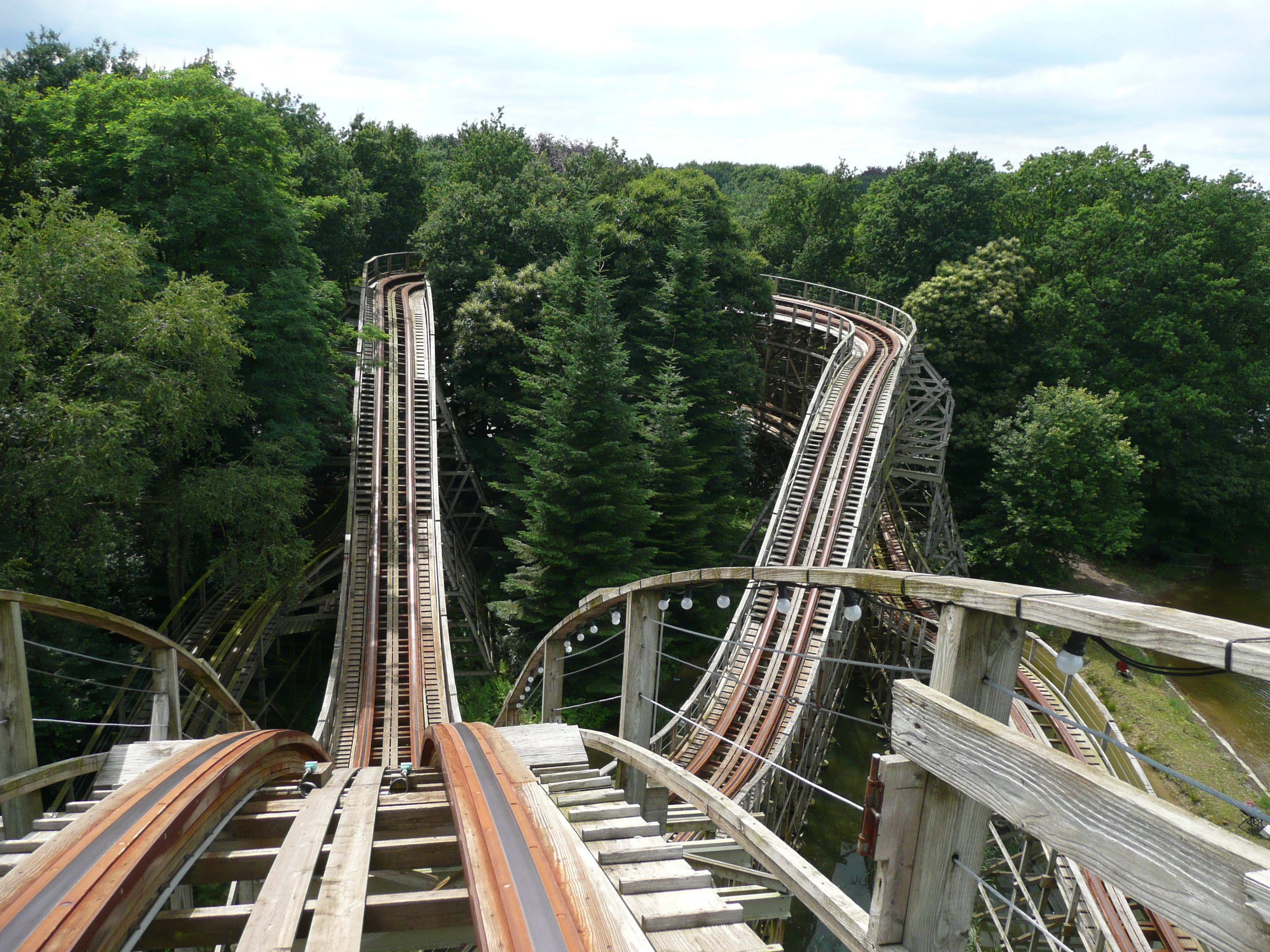 View of a wooden roller coaster track among green trees from the viewpoint of a person riding in the front car.