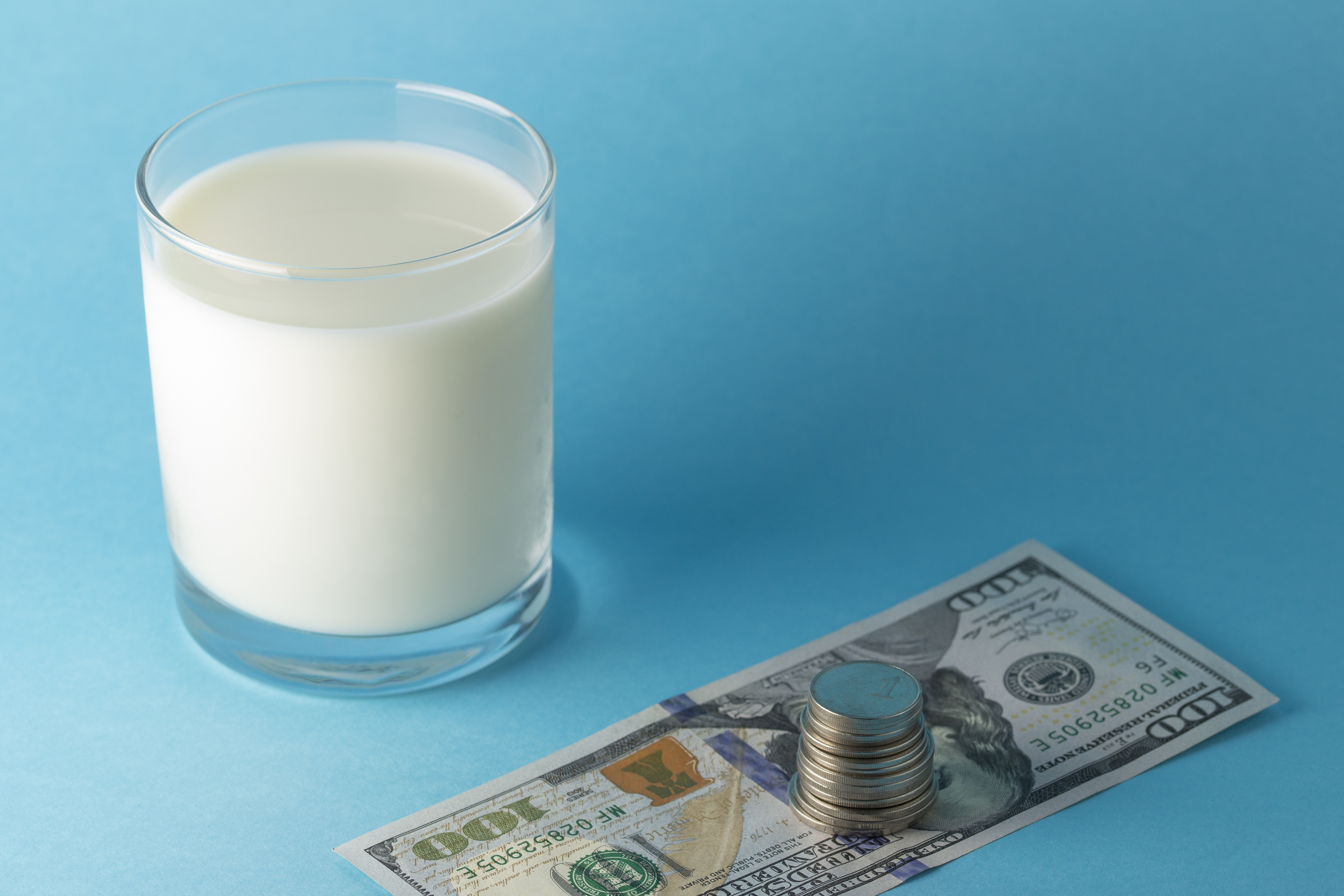 Glass of milk on a light blue surface, next to a U.S. $100 pill with a stack of coins set atop it.