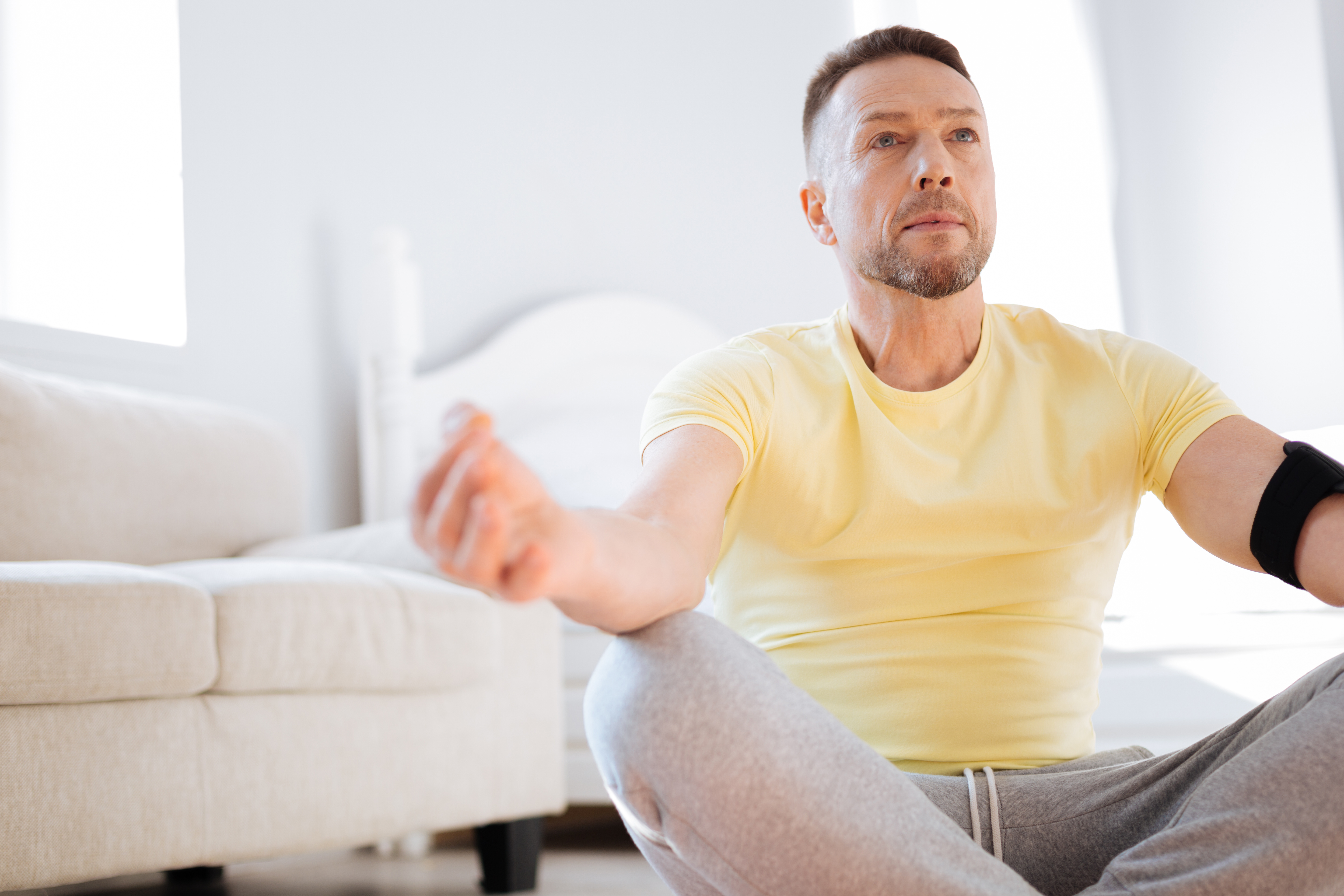 Photo of a man sitting cross-legged on the floor in front of a white sofa. He wears a yellow t-shirt and gray sweatpants.  His outstretched arms rest on his knees as he gazes forward.