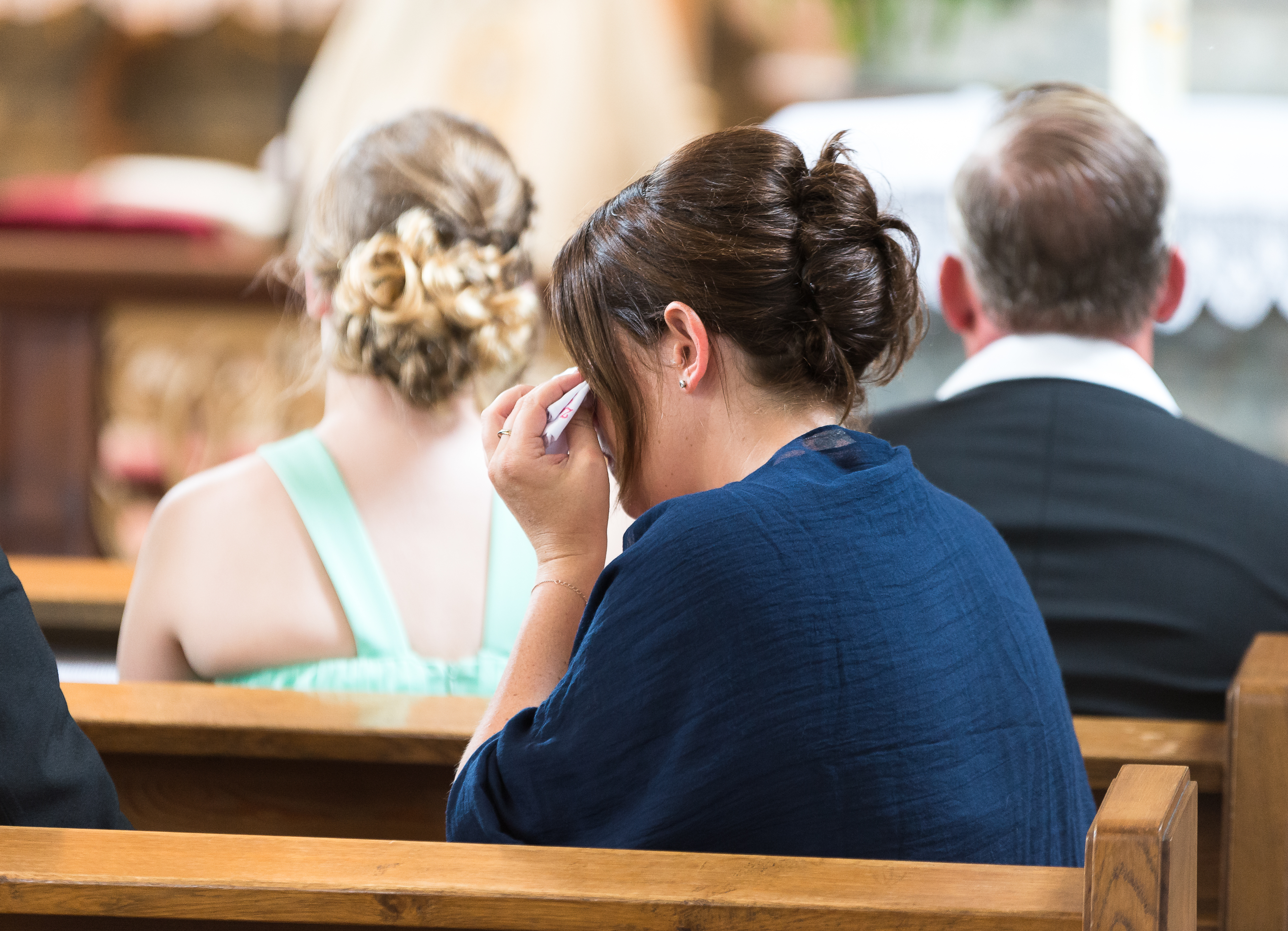 Woman wiping her eyes with tissue, sitting in church pew behind two other people.