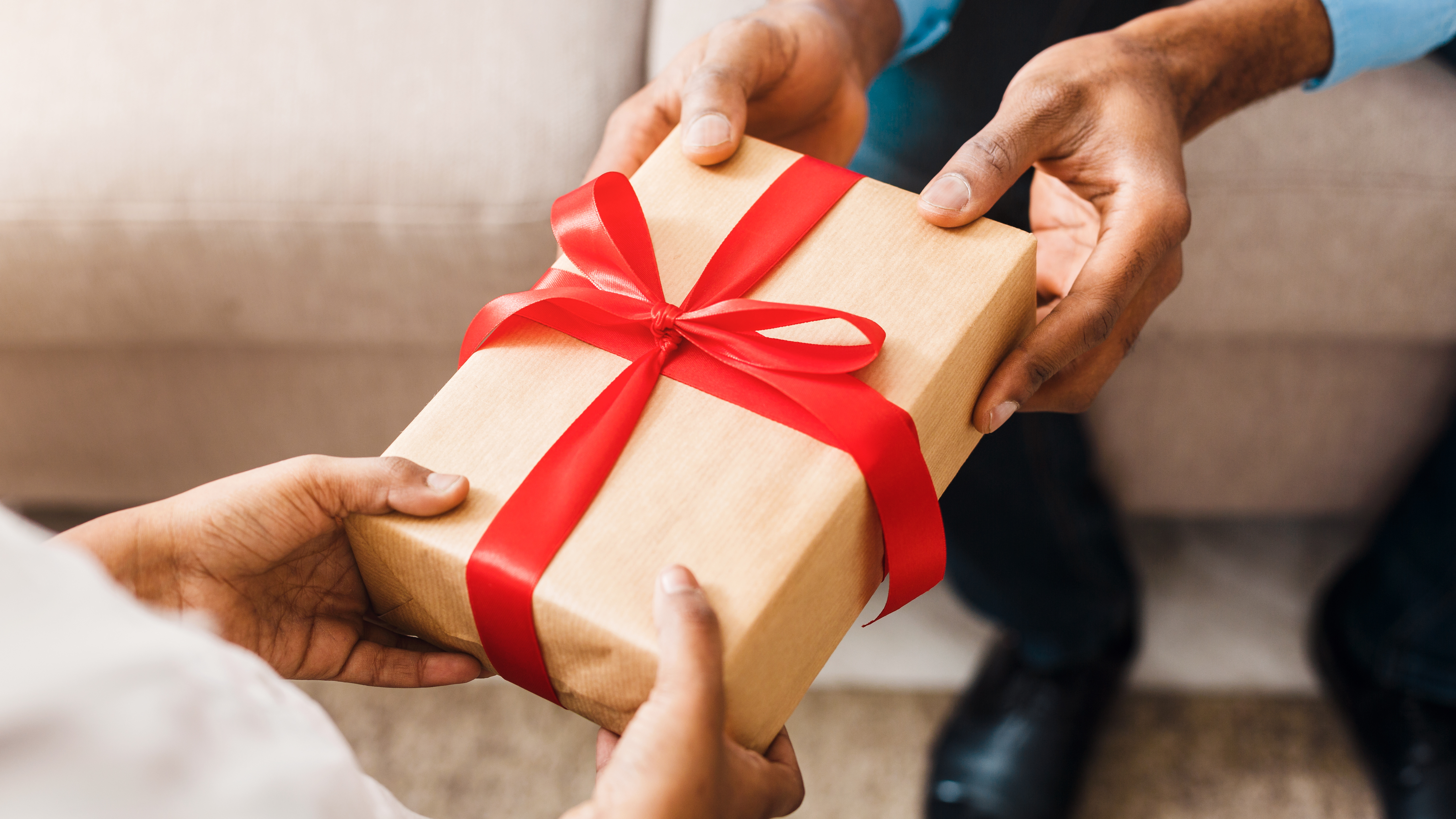 Package wrapped in brown paper with red ribbon tied around it in a bow; hands of one person placing package in another person's hands.