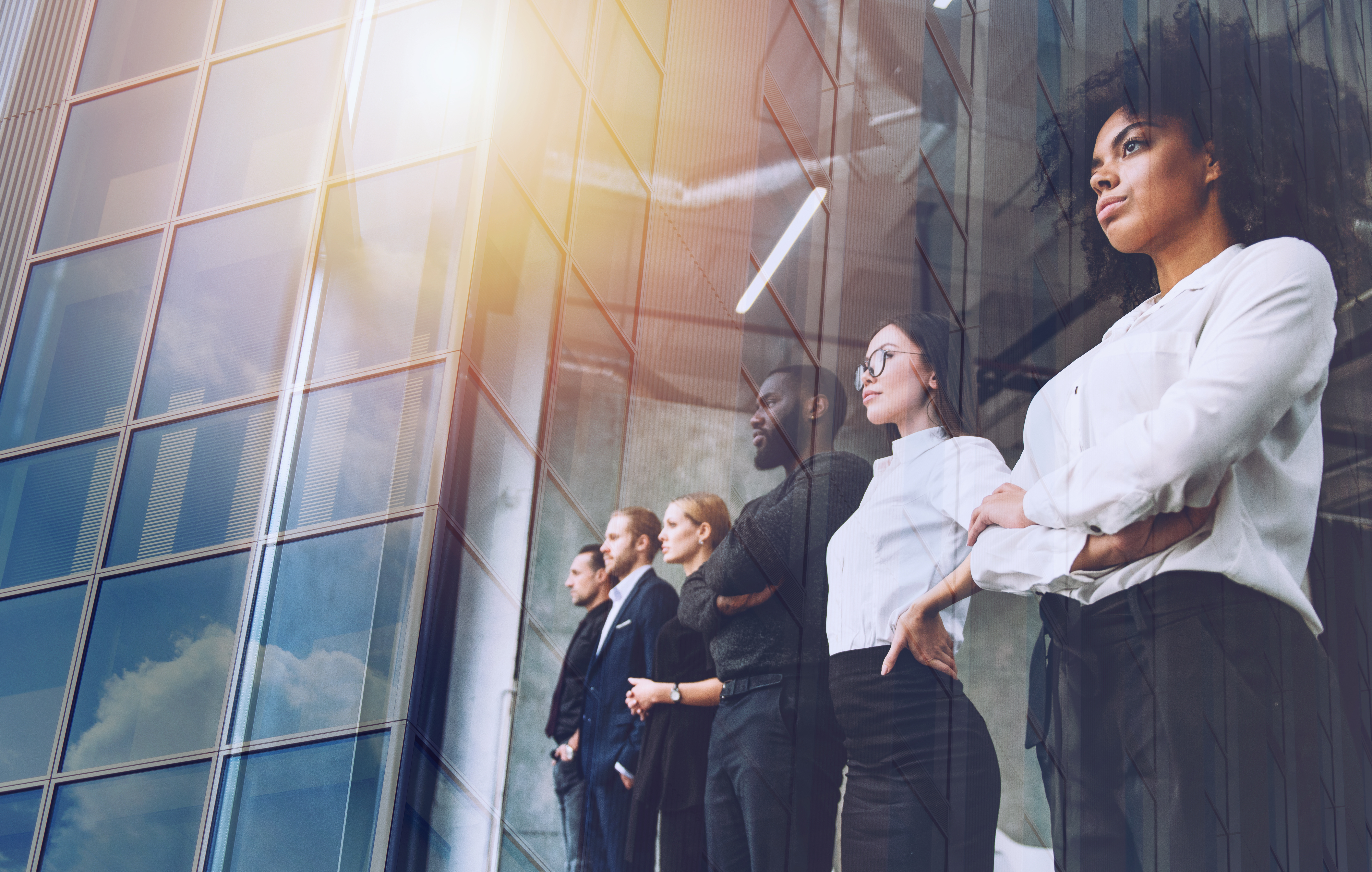 Photo: at right, a diverse group of young professionals stands in a row, all looking out through a large glass wall. Nearby is a large glass office building, which is reflected in their own window.