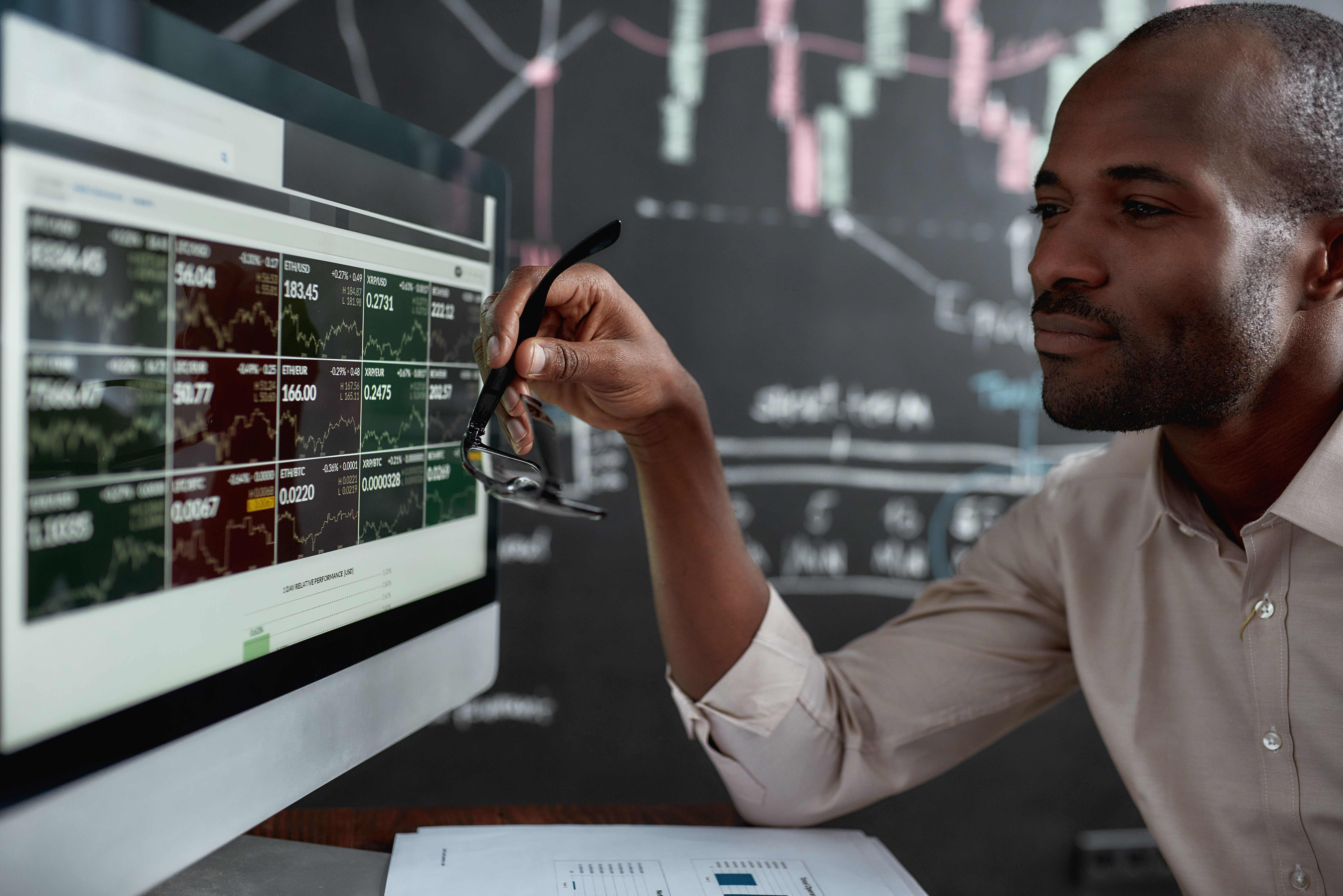 Image of a person wearing a button-down shirt, holding eyeglasses, looking at 15 charts on a computer screen, with printed charts on the desk and a large chart drawn on a blackboard in the background.