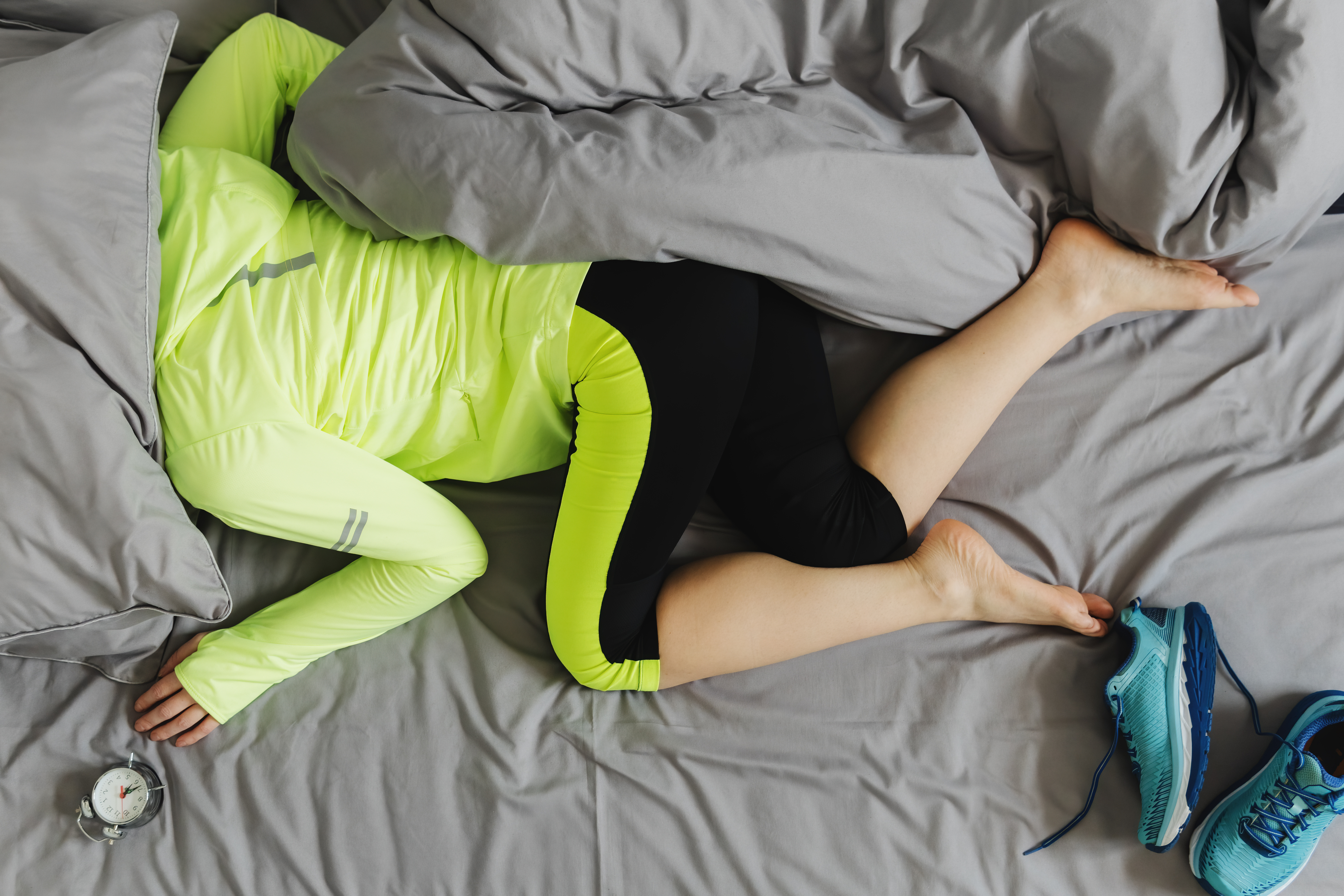 Woman in neon green hooded sweatshirt and black workout pants with matching green stripe, lying on an unmade bed with gray linens, head hidden under pillow.