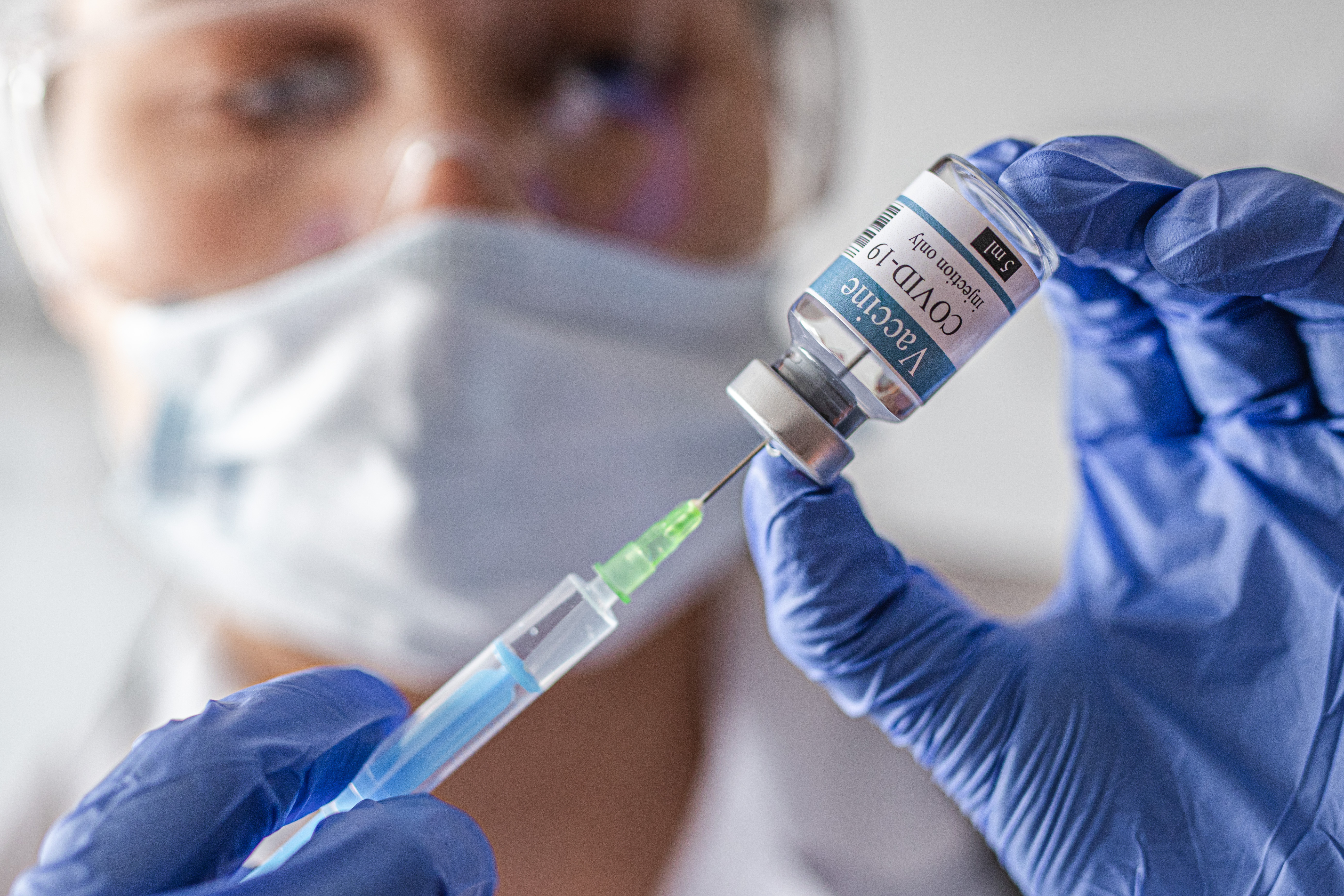 Image of person wearing a surgical mask, safety glasses, and plastic gloves drawing liquid into a syringe from an ampoule labeled "Vaccine COVID-19."