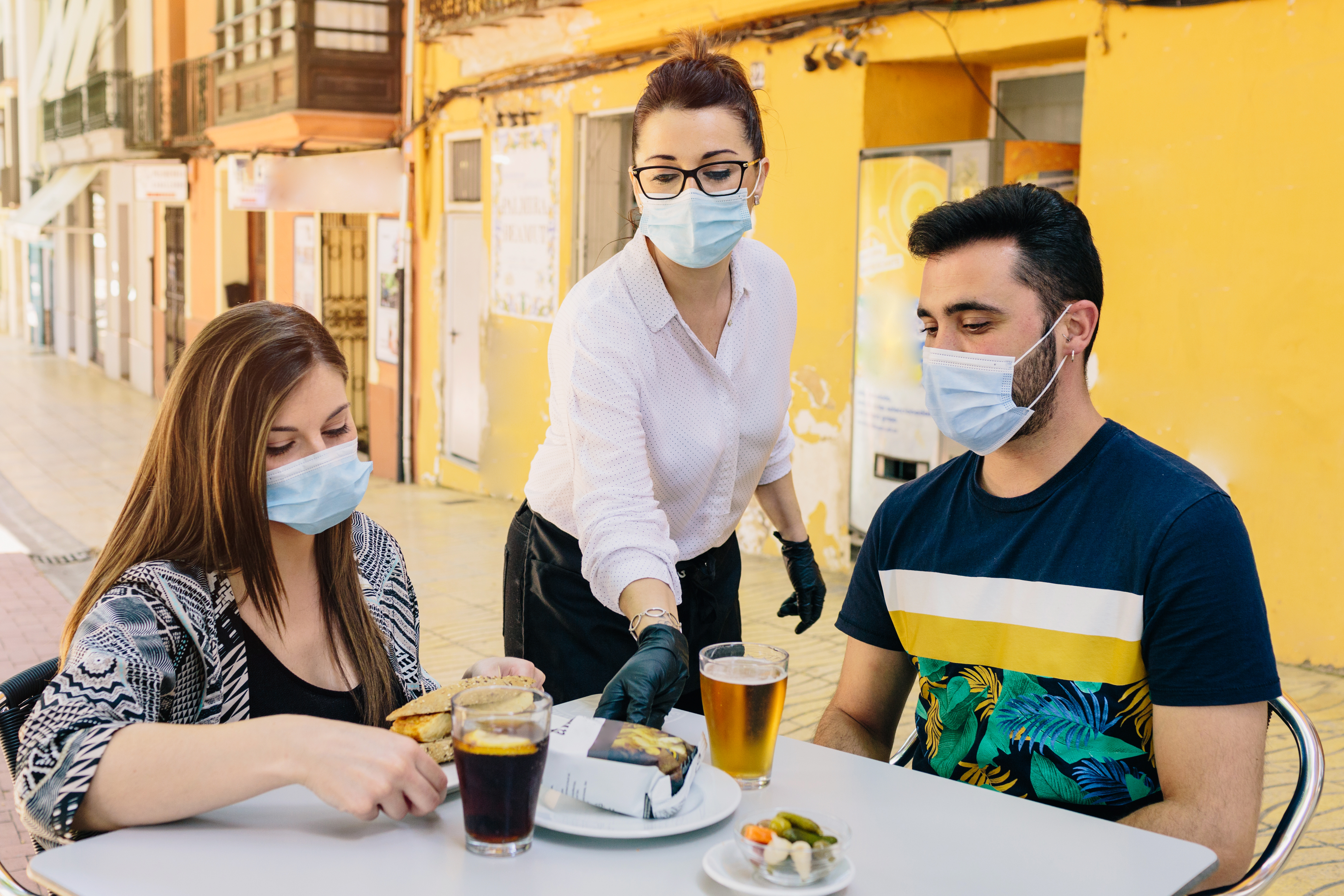 A waitress sets a plate between two customers seated at an outdoor table.  All three people wear surgical masks; the waitress also wears gloves.