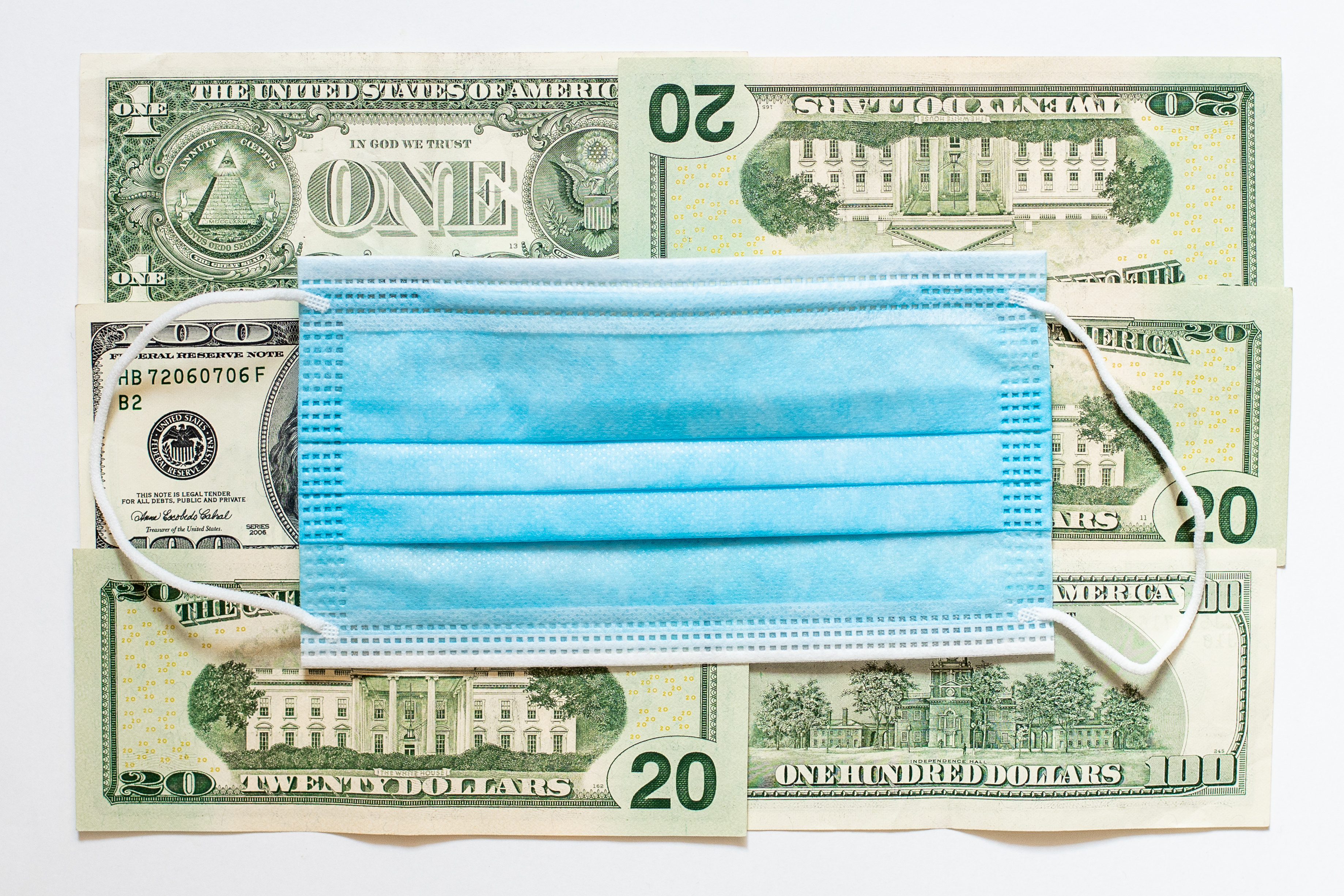Photo of neatly arranged U.S. paper currency with a blue surgical mask placed on top of it.