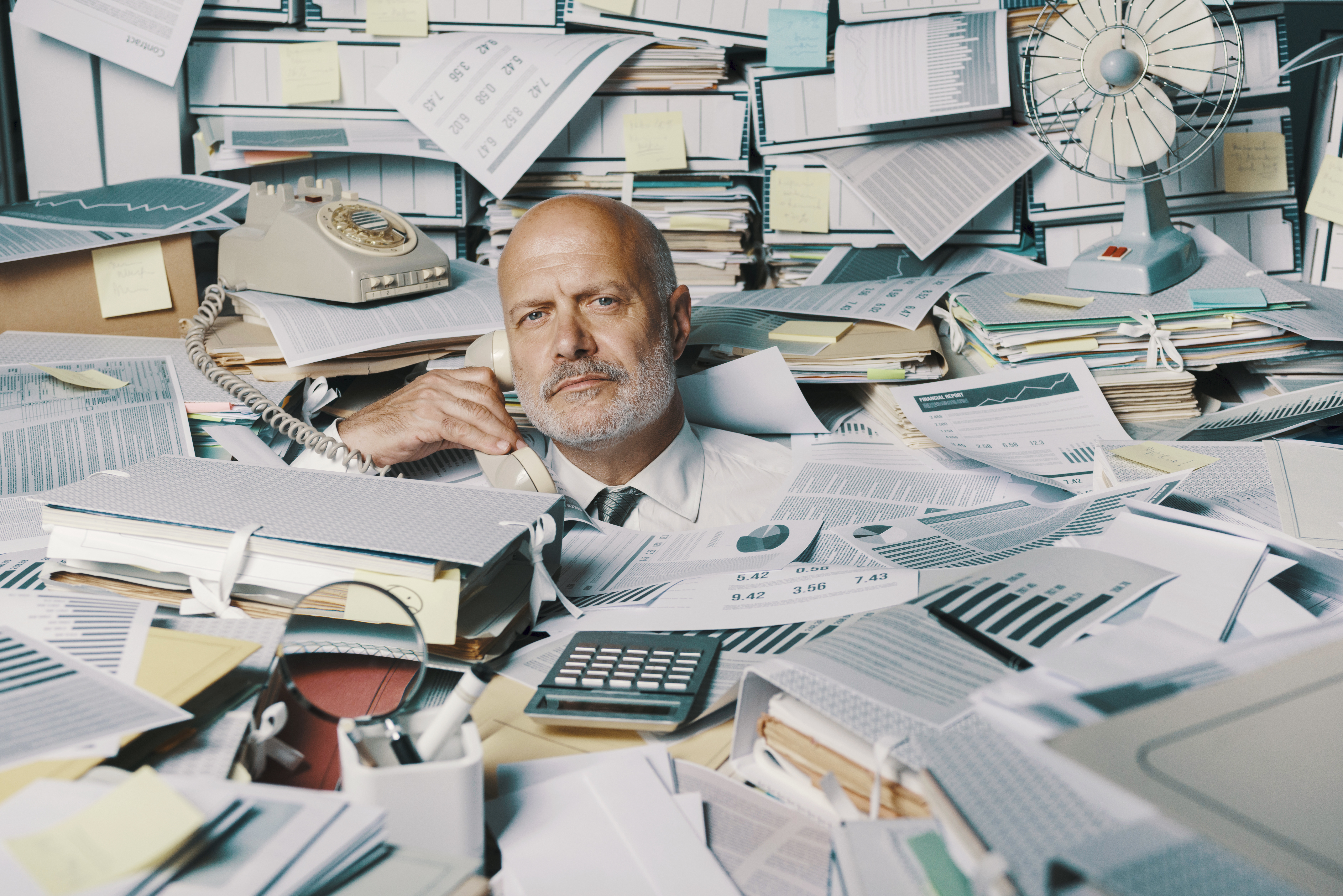 Photo of a businessman looking at the camera, holding the receiver of a rotary phone, buried up to his neck in papers, books, and other office paraphernalia.