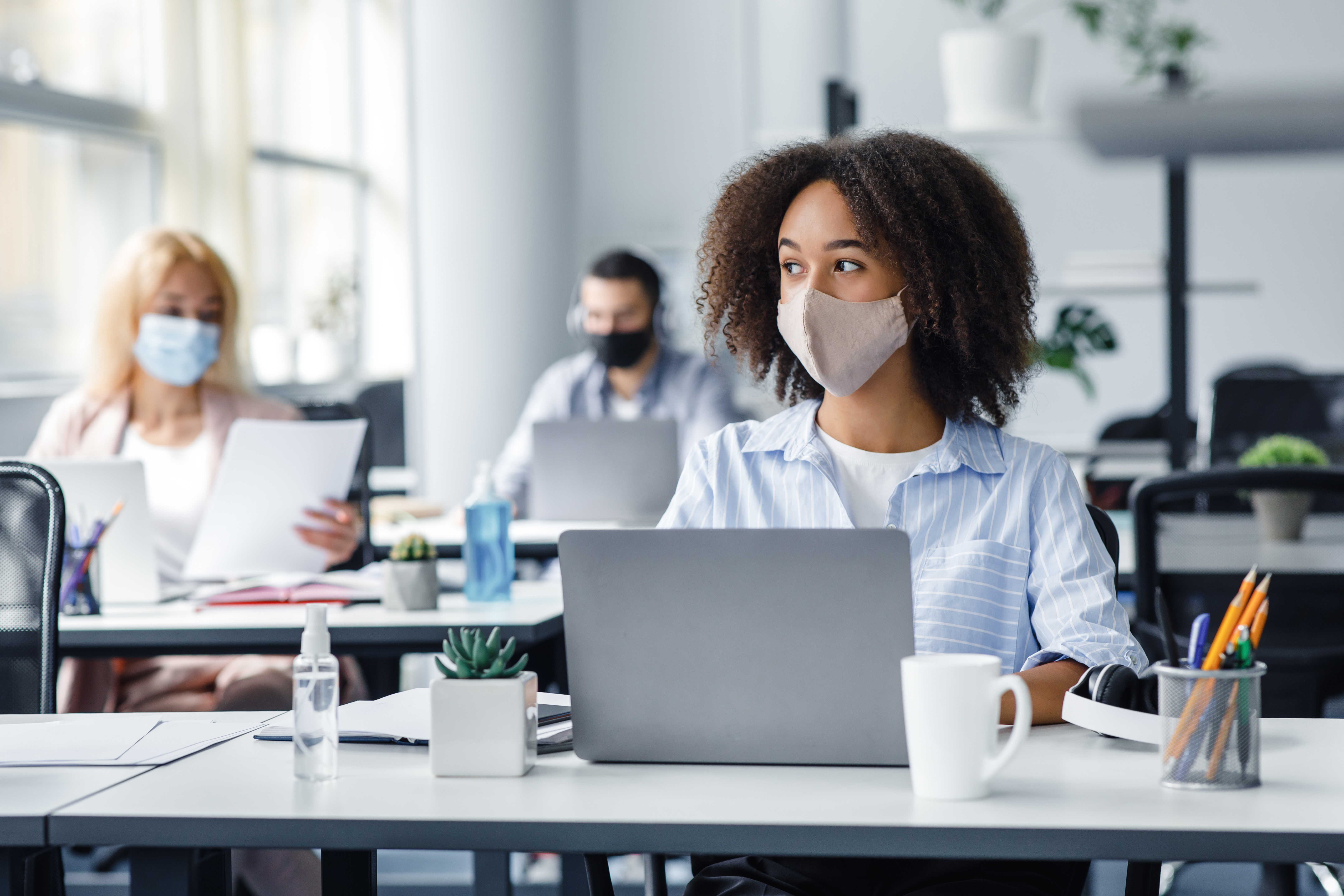 Photo of an office with three people wearing face masks, seated at separate desks at a distance from each other.  The woman closest to the camera looks off to the side into the distance.