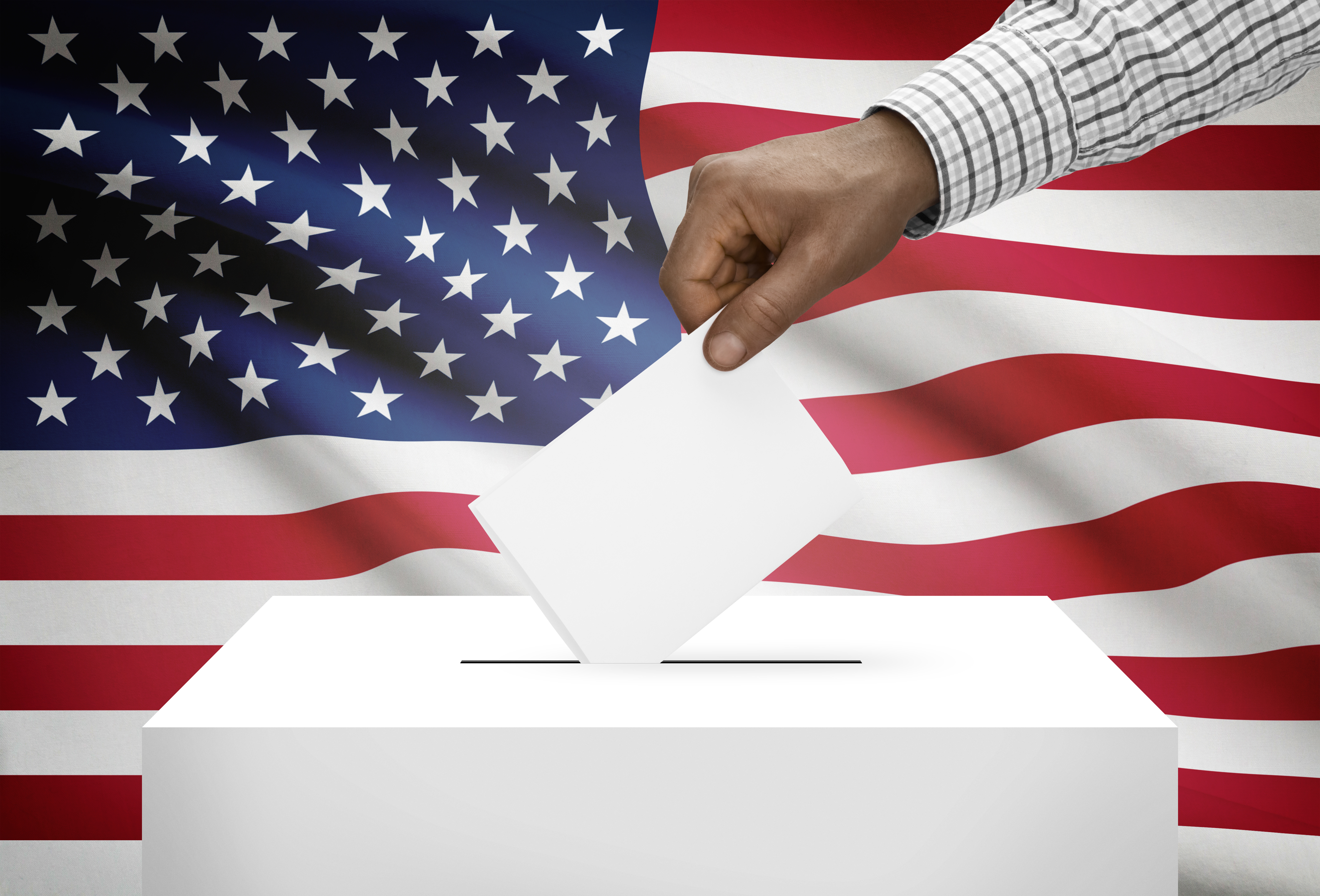 Hand and arm of person placing white paper ballot into white ballot box with American flag in background.
