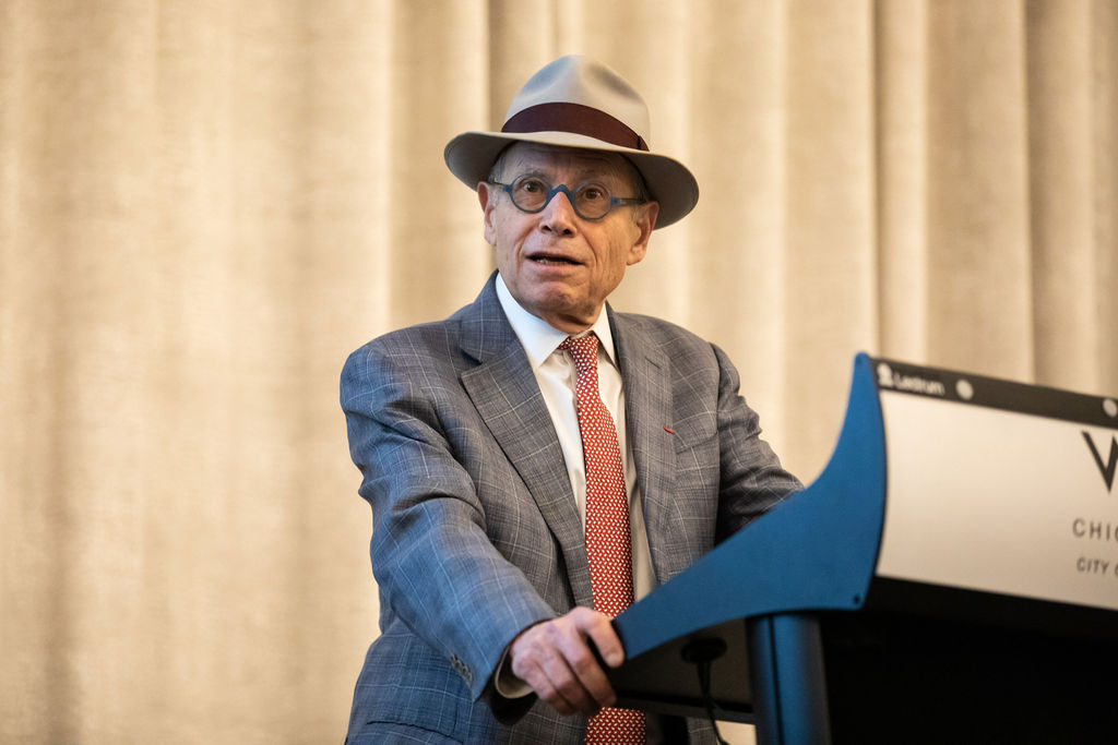 A man in a gray suit, red tie, and tan fedora speaks at a podium in front of a tan curtain.