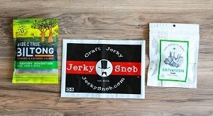 Three items on light wooden table: green package of Biltong jerky; card with red, white, and black Jerky Snob logo; and white package of Southwestern jerky.